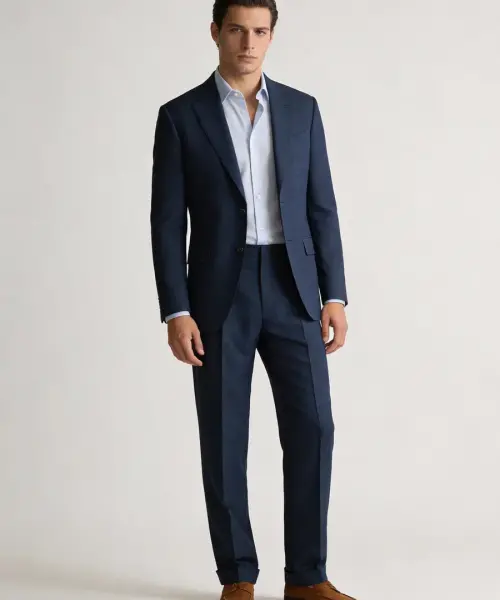 Homme portant un costume bleu marine à coupe droite avec chemise bleu clair sans cravate, pantalon assorti et mocassins marron, sur fond neutre.