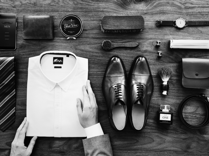 Flat lay en noir et blanc : chemise blanche pliée, cravate rayée, chaussures en cuir, montre, portefeuille, ceinture, parfum, boutons de manchette et accessoires d’entretien sur table en bois.